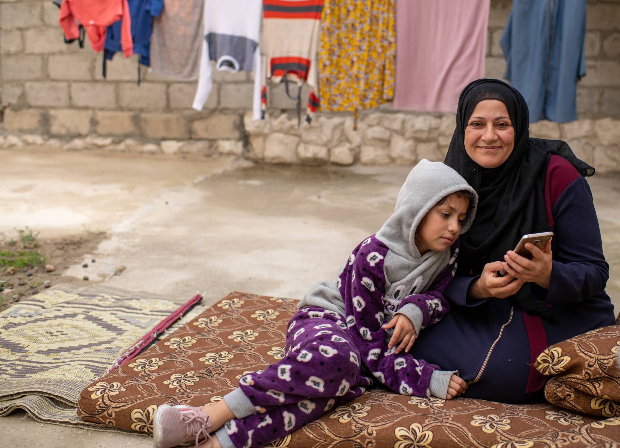 Nawal smiles at the camera while her daughter looks at a smart phone Nawal is holding. 