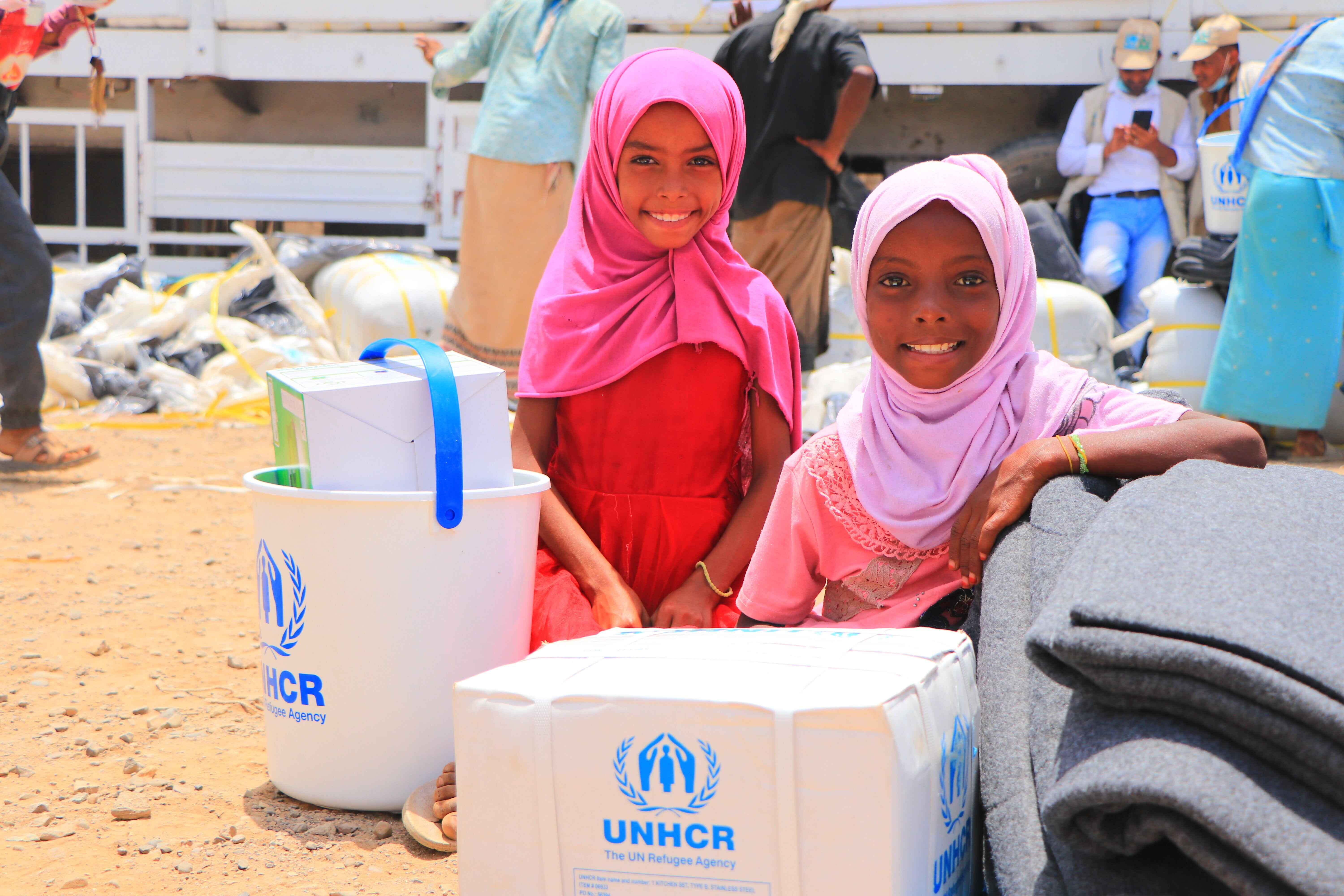 Two refugee girls smile surrounded by UNHCR supplies