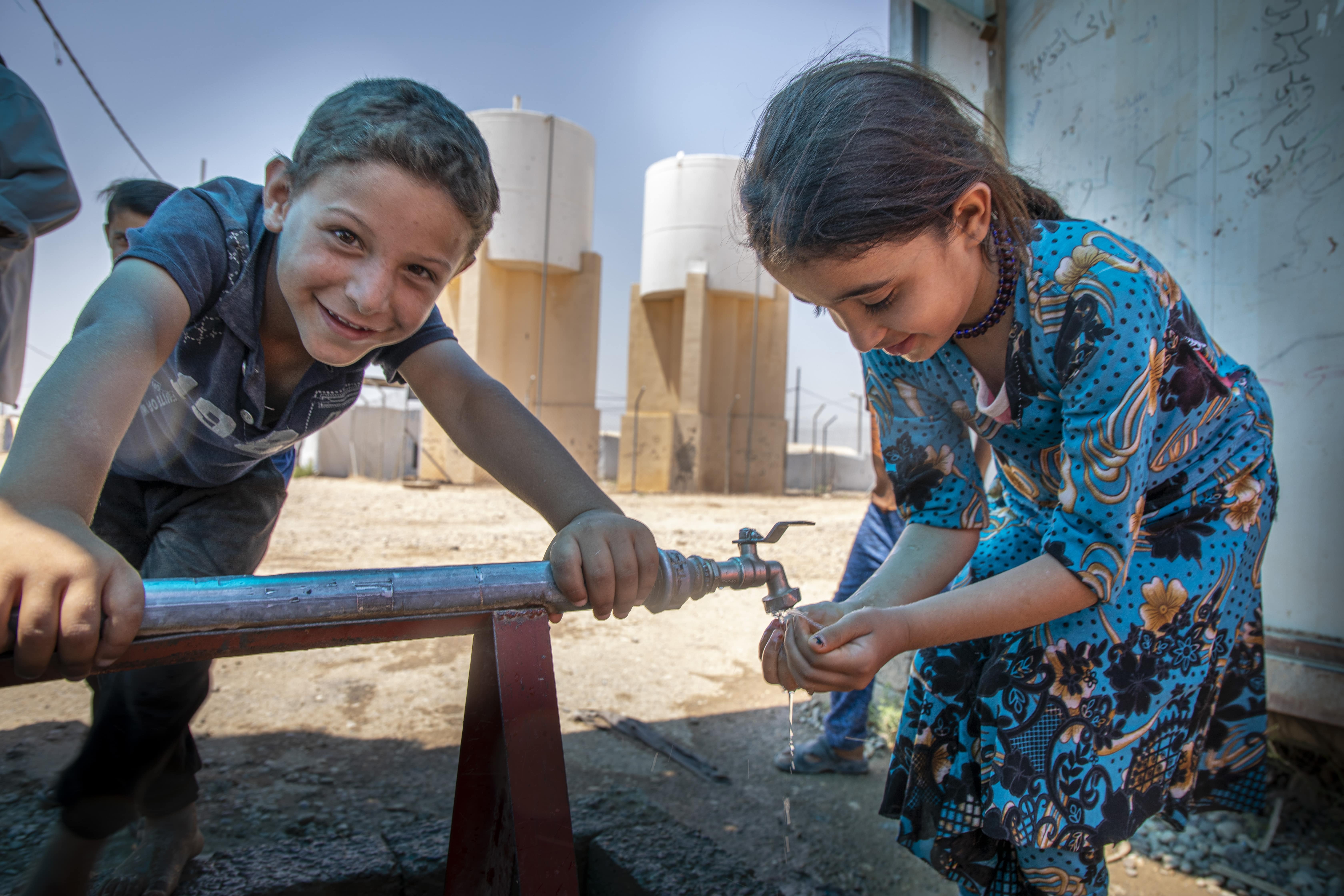 A boy and a girl in a refugee camp in Syria. The girl is having some water from the communal water tap. Providing potable water and raising awareness among children about the importance of hand washing is essential for preventing the spread of COVID-19.